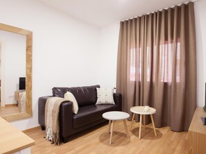 A modern living room with a dark leather sofa, two small round tables, a large mirror, and a view through sheer curtains.