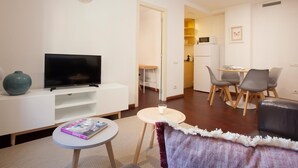 Una sala de estar moderna con un televisor de pantalla plana sobre un mueble blanco, una mesita redonda con una revista, un sofá y un comedor con una mesa y sillas.