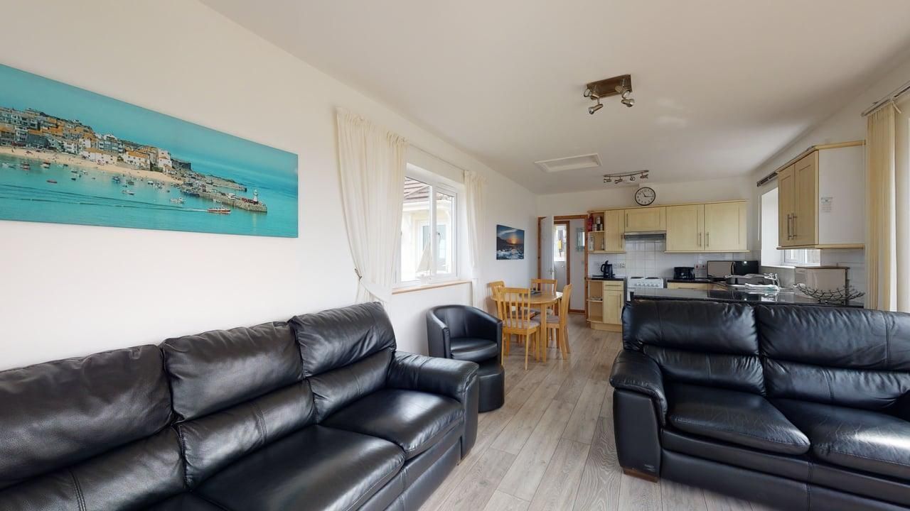 A living room with two black leather sofas, a dining table with chairs, a kitchen area with cabinets and appliances, and a painting of a coastal scene on the wall.