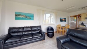 Ein Wohnzimmer mit zwei schwarzen Ledersofas, einem Esstisch mit Stühlen, einer Küchenzeile und einem Fenster mit Vorhängen.