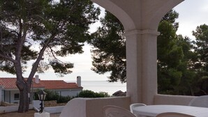 Eine Terrasse im Freien mit weißen Kunststoffstühlen und einem Tisch, mit Blick auf ein Gewässer und ein Gebäude mit roten Ziegeldach.