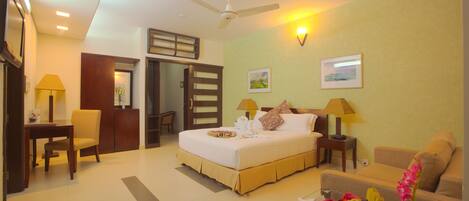 A hotel room with a bed, a desk, a chair, a sofa, a glass coffee table, a fruit basket, and a vase with flowers.