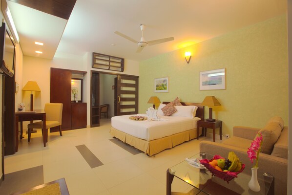 A hotel room with a bed, a desk, a chair, a sofa, a glass coffee table, a fruit basket, and a vase with flowers.