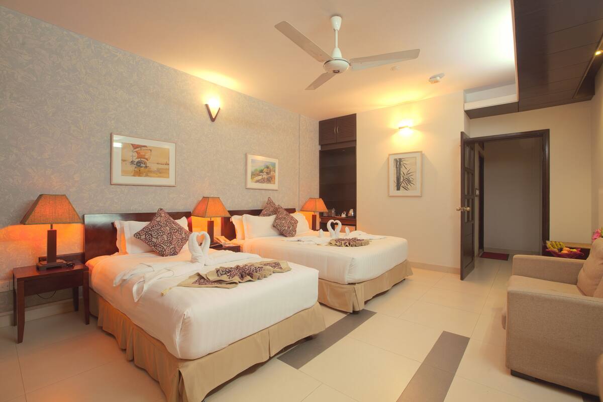 A hotel room with two beds, a ceiling fan, wall-mounted lamps, and a door leading to another room.