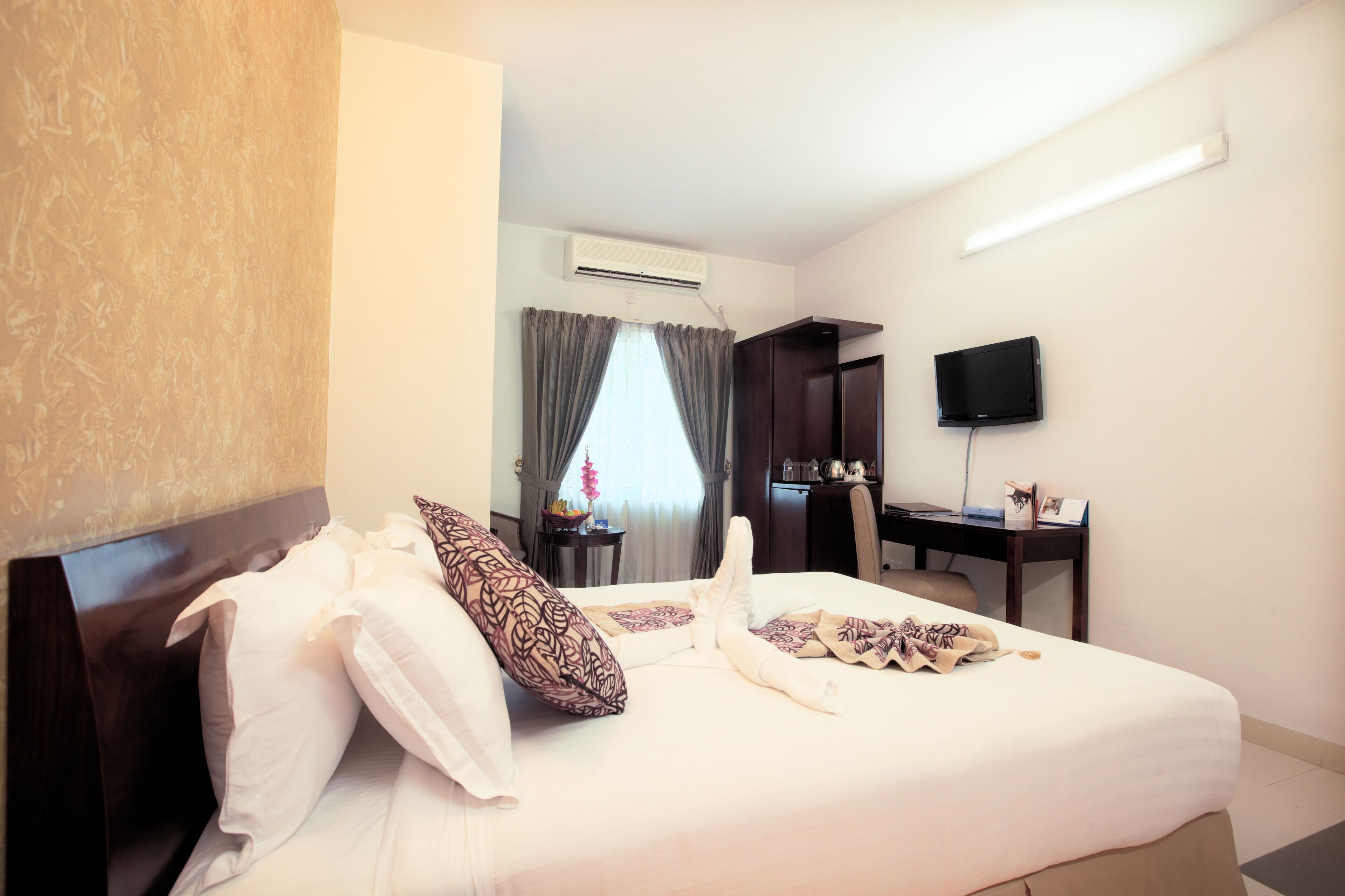 A hotel room with a neatly made bed, a desk with a chair, a flat-screen TV mounted on the wall, and an air conditioning unit.