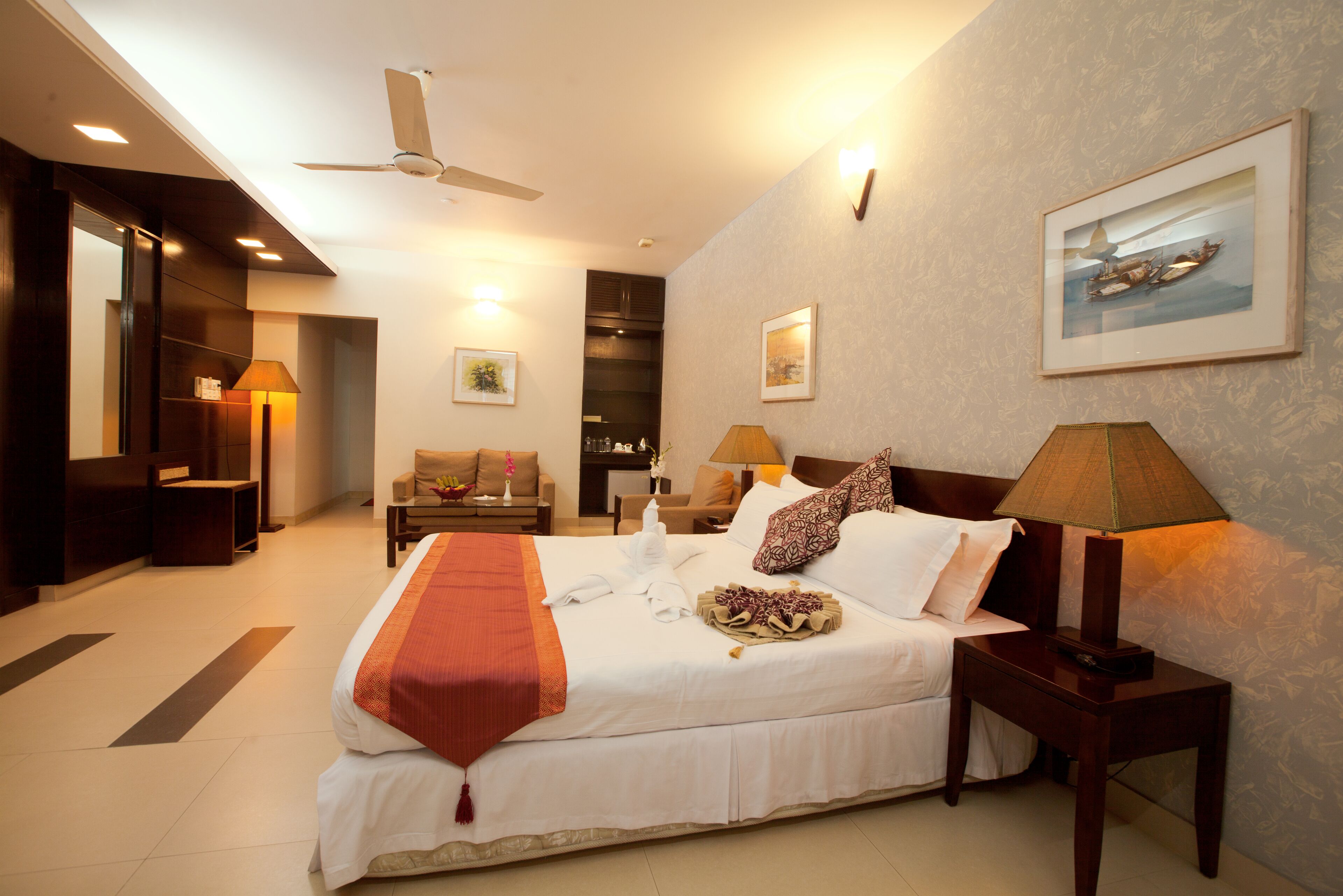 A hotel room with a large bed, a ceiling fan, a nightstand with a lamp, a sofa, and a painting on the wall.