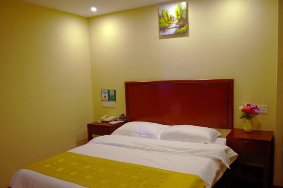 Hotelkamer met een bed, een houten hoofdbord, een nachtkastje met telefoon, een vaas met bloemen en een schilderij aan de muur.