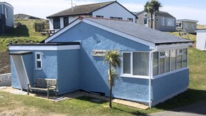 A single-story house with a sloped roof, a small porch with a bench, and a palm tree in front. - D34 Homeleigh, Riviere Towans (Hayle)