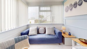 A cozy room with a blue sofa, a wooden side table with a lamp, and a view of the outdoors through large windows with blinds. - D34 Homeleigh, Riviere Towans (Hayle)