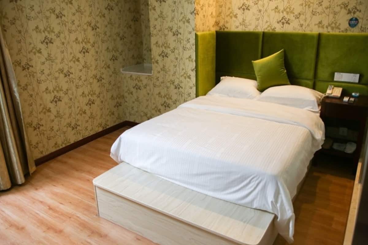 Un lit bien fait, avec du linge de lit blanc et une tête de lit verte, un banc en bois au pied du lit, et une table de chevet avec un téléphone et une petite table.