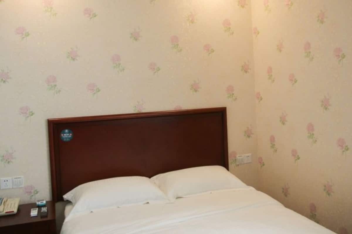 Un lit simple avec une tête de lit en bois, dans une pièce dont les murs sont recouverts d’un papier peint à motifs floraux.
