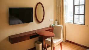 Une chambre d’hôtel équipée d’un téléviseur à écran plat fixé au mur, d’une table de chevet en bois, d’une chaise blanche, d’une petite poubelle et d’une fenêtre avec des rideaux.