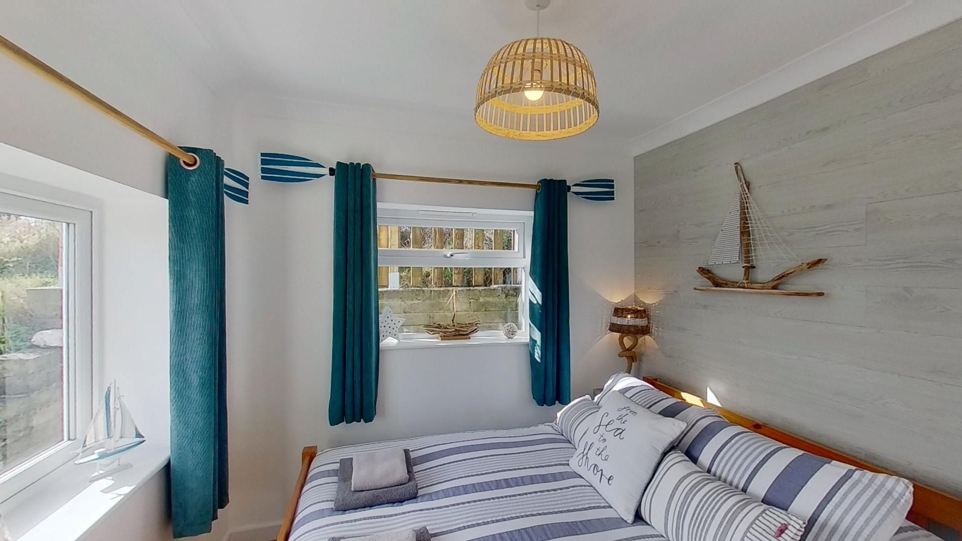 A bedroom with a bed, a window with curtains, a wall-mounted shelf with a sailboat model, and a hanging light fixture.