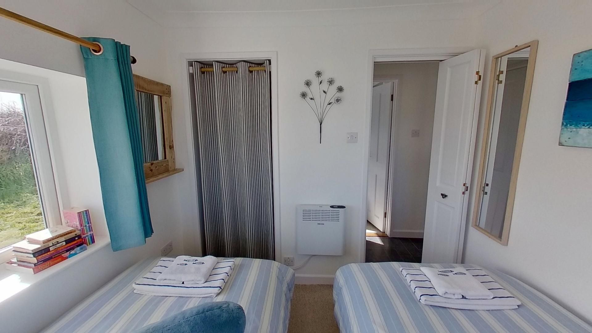 A small, clean room with two single beds, a window with curtains, a mirror, and a wall-mounted air conditioner.