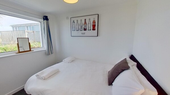 A bedroom with a bed, a window with curtains, a framed picture of Coca-Cola bottles, and a view of a fence outside.