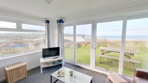A spacious living room with a large TV on a white stand, a glass coffee table, and a view of the beach and fields through large windows. - B25 Trezaren, Riviere Towans (Hayle)