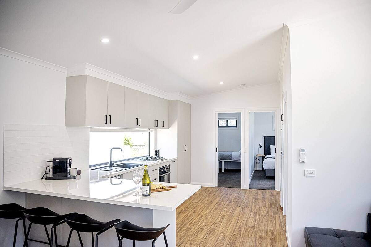 Een moderne keuken met witte kasten, een groot eiland met barkrukken, en uitzicht op een slaapkamer.