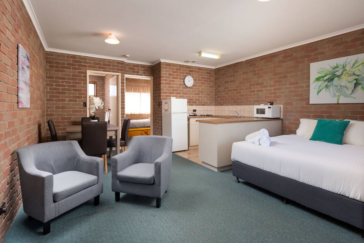 Een compacte hotelkamer met een bakstenen muur, een kitchenette, een bed, twee fauteuils en een kleine tafel.