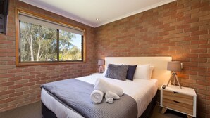 Een slaapkamer met een bakstenen muur, een groot raam, een bed met een grijze deken, een nachtkastje met een lamp en een nachtkastje.