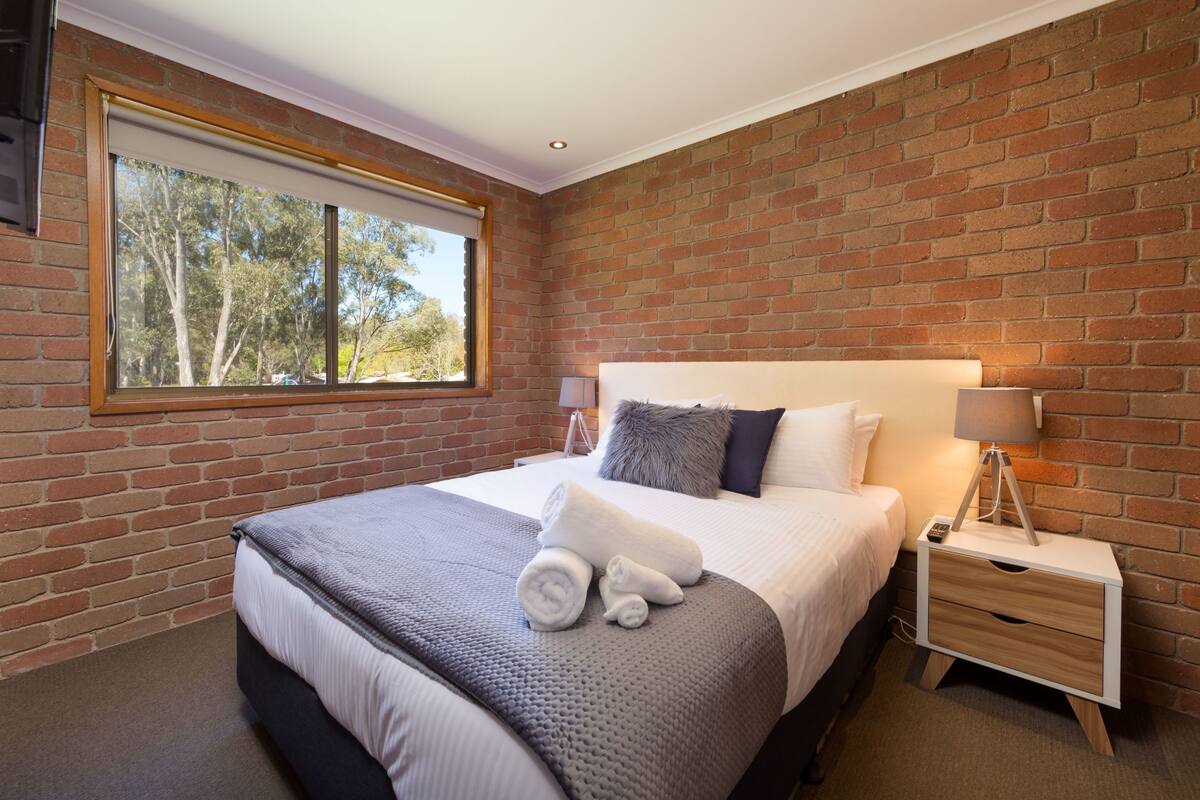 Een slaapkamer met een bakstenen muur, een groot raam, een bed met een grijze deken, een nachtkastje met een lamp en een nachtkastje.