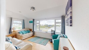 A spacious living room with a large window, a sofa, a teal armchair, and a TV.