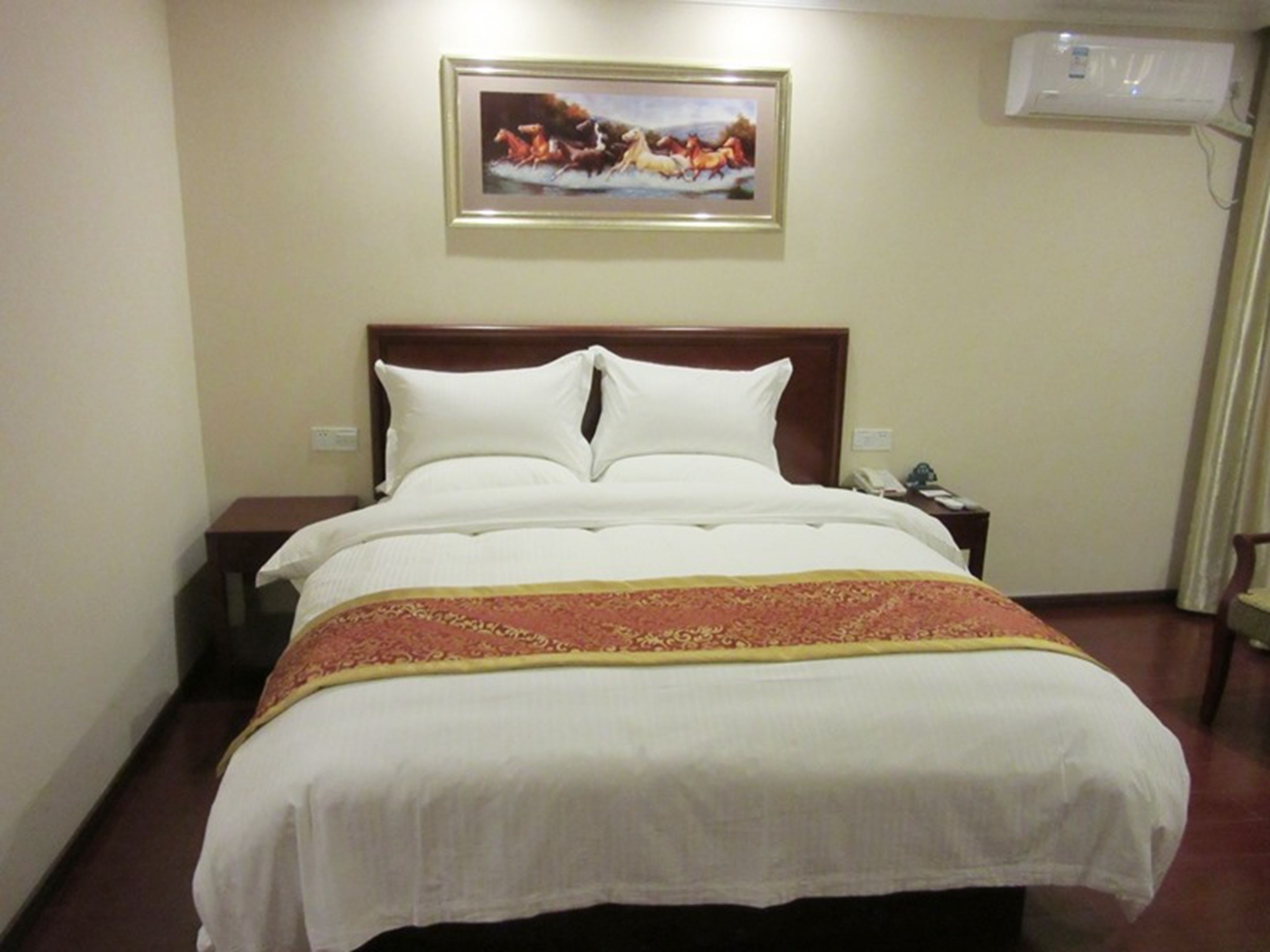 A hotel room with a large bed, a nightstand, a framed picture on the wall, and an air conditioning unit.