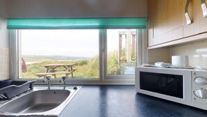 A kitchen with a large window, a sink, a microwave, and wooden cabinets. - C37 Jupiter, Riviere Towans (Hayle)