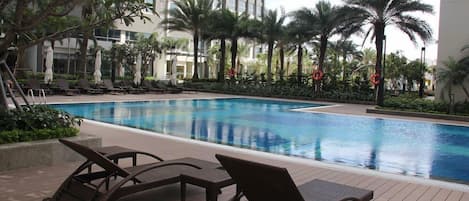 Una piscina con sillones de descanso y sombrillas, rodeada de palmeras y un edificio moderno al fondo.