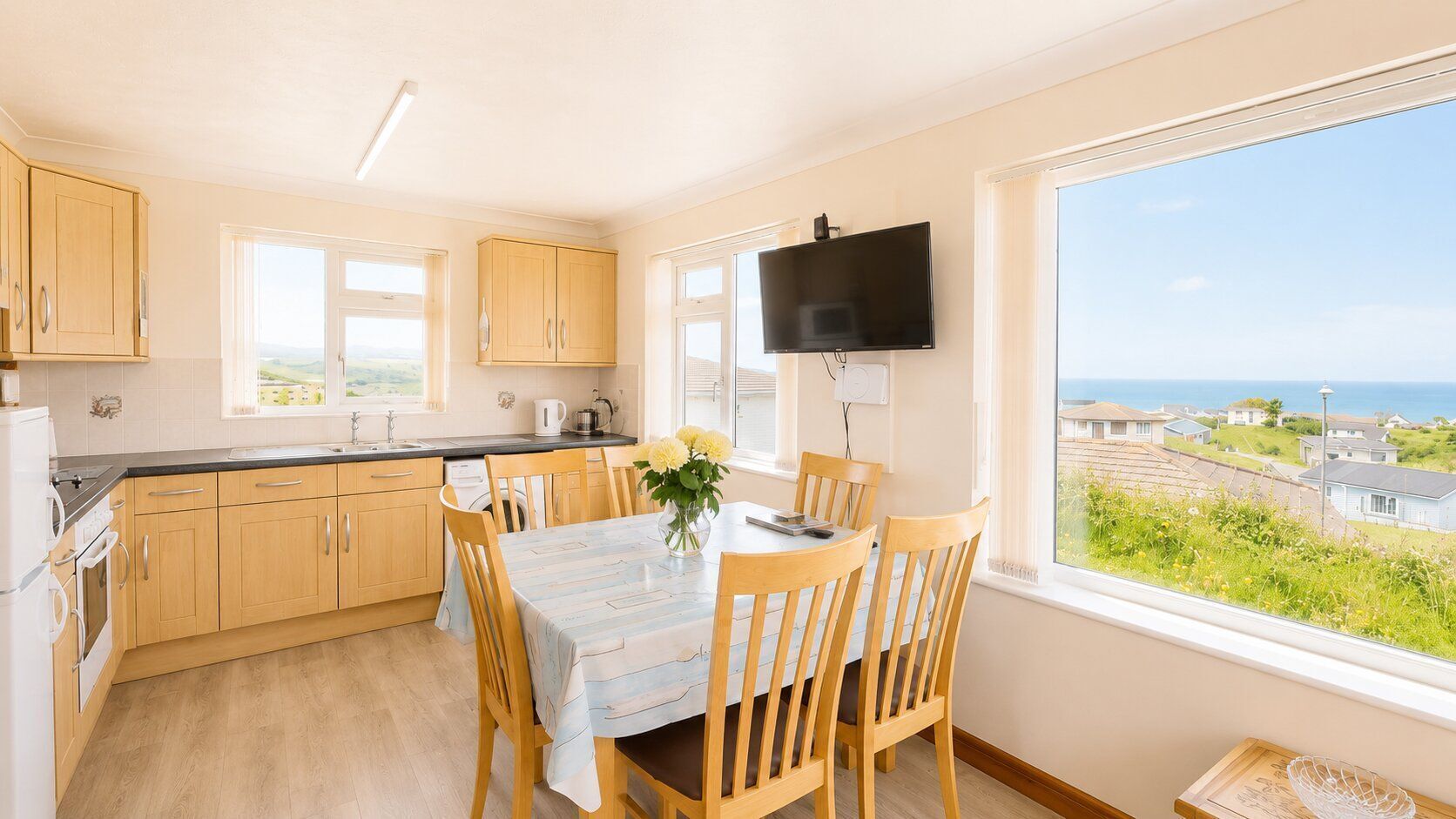 A kitchen with wooden cabinets, a dining table with chairs, a TV mounted on the wall, and a window with a view of the sea.