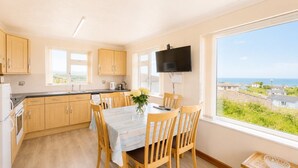 Una cocina con armarios de madera, una mesa de comedor con sillas, un televisor montado en la pared y una ventana con vista al mar.