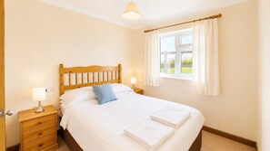 A bedroom with a wooden bed, white bedding, a nightstand with a lamp, and a window with curtains.