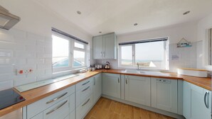 A modern kitchen with light blue cabinets, wooden countertops, and large windows offering views of the sea. - C20 Y Worry, Riviere Towans (Hayle)
