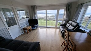 A living room with a black sofa, a wooden dining table, a TV, and large sliding glass doors to a garden. - C20 Y Worry, Riviere Towans (Hayle)