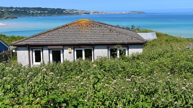 A building with a tiled roof, surrounded by lush greenery and overlooking a coastal area with clear blue waters. - C20 Y Worry, Riviere Towans (Hayle)