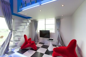 Ein modernes Zimmer mit Schachbrettmuster-Boden, einem Fernseher, roten Stühlen, einem Glastisch und einer Treppe mit Regalen.