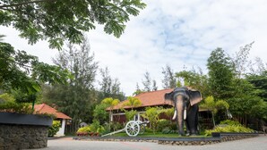 Une grande statue d’éléphant, avec un attelage, se trouve dans un espace aménagé avec des arbres et un bâtiment doté d’un toit rouge.