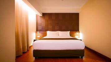A hotel room with a large bed, wooden headboard, bedside lamps, and a curtain on the left.