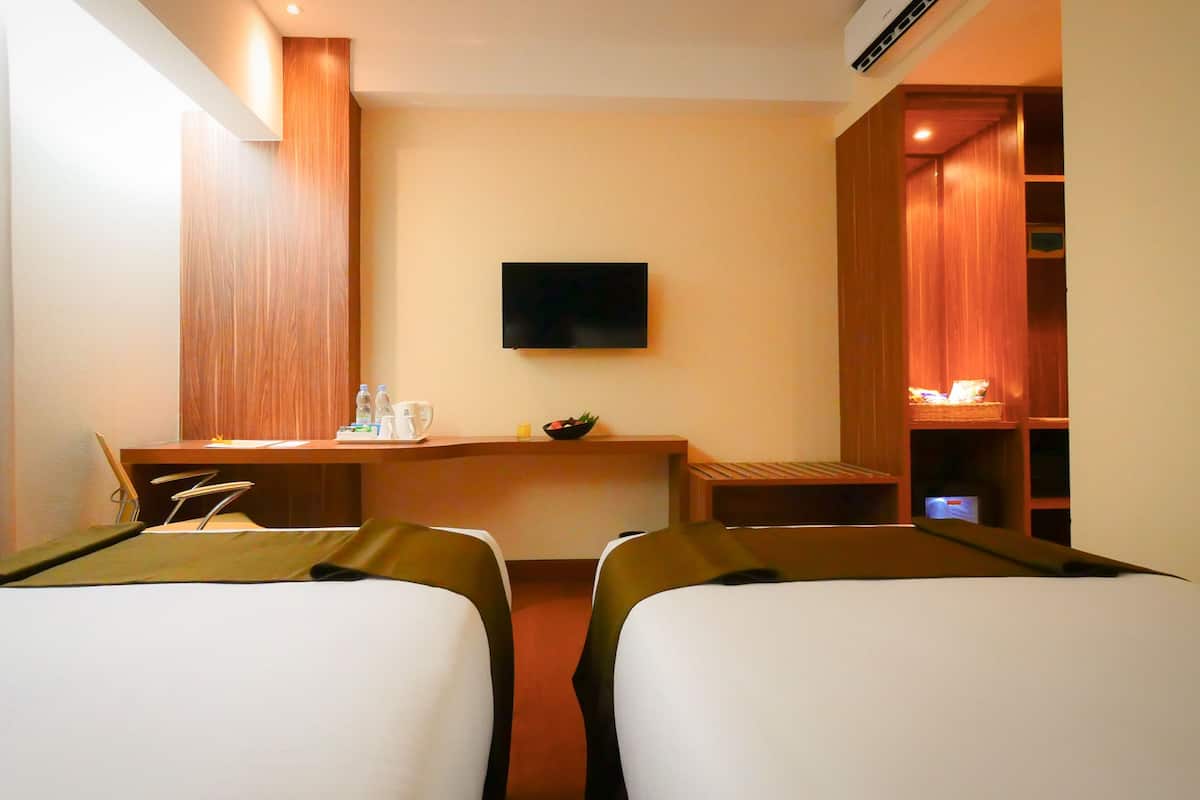 Une chambre d’hôtel avec deux lits, un bureau doté d’un téléviseur à écran plat, une petite table avec un panier de fruits et une bibliothèque.