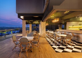 An outdoor dining area with checkered flooring and patterned chairs, leading to a bar area with a counter and pendant lighting.