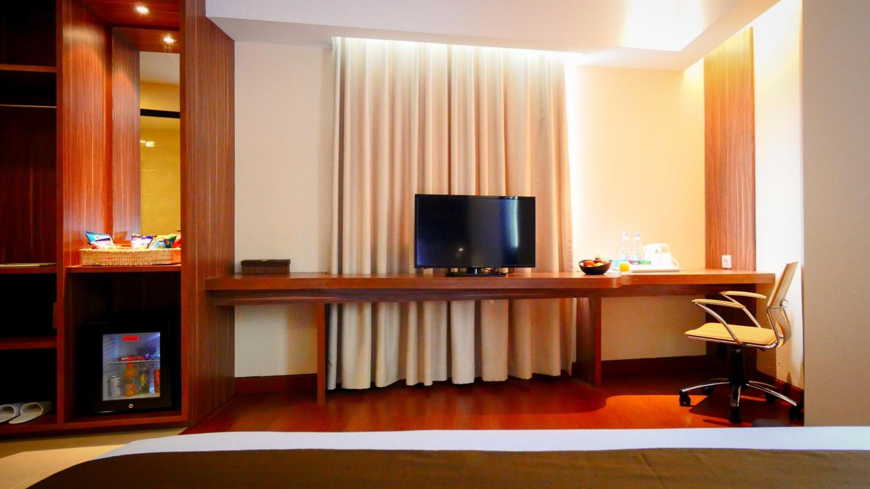 A hotel room with a wooden desk, a television, a chair, and a shelf with snacks.