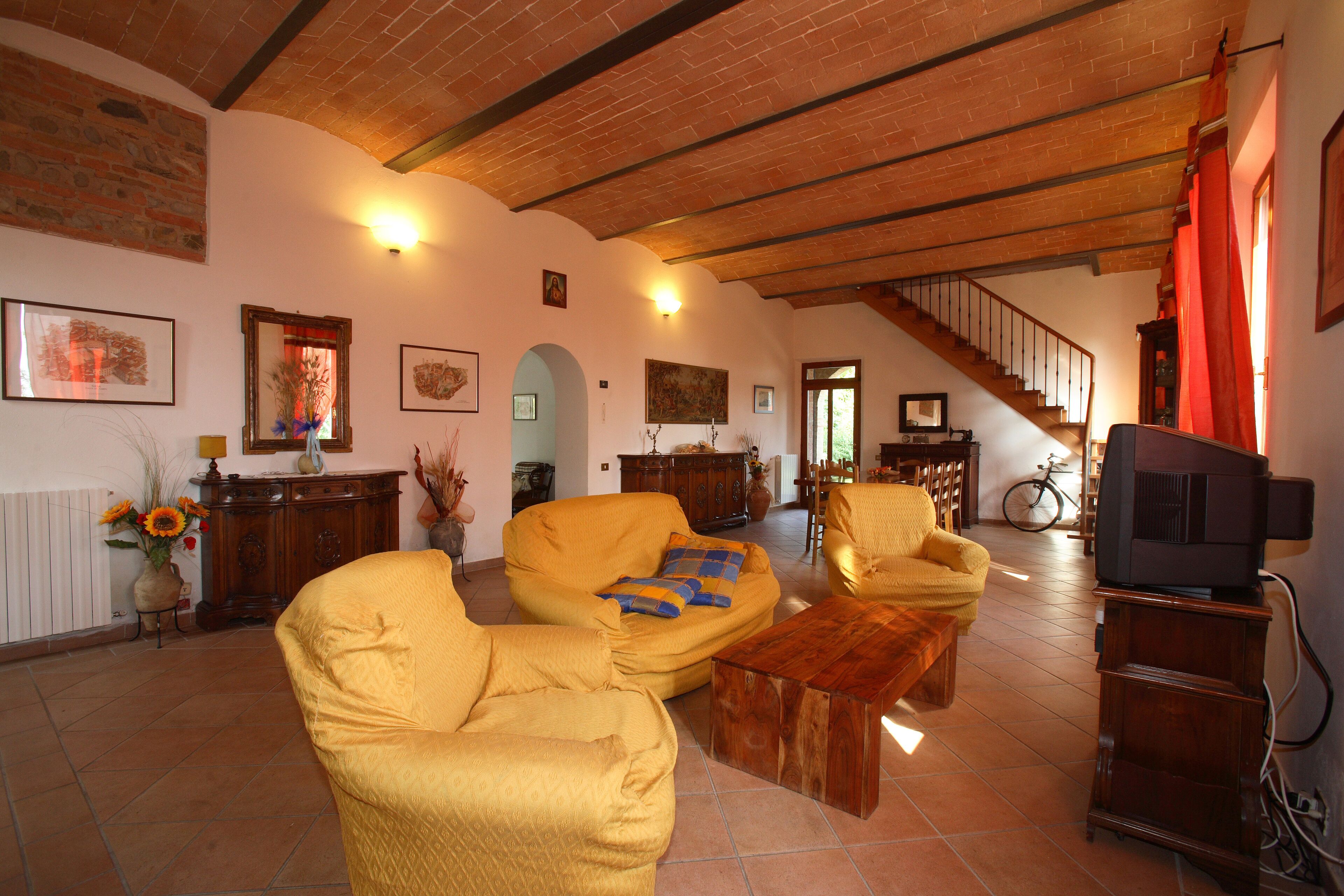 A spacious living room with yellow armchairs, a wooden coffee table, and a television. A staircase leads to an upper level.