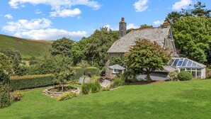 Une maison en pierre avec un jardin, une serre et une colline en arrière-plan.