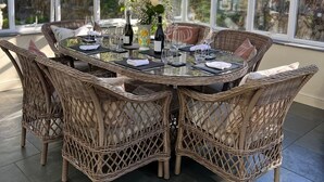 Une table ronde en verre, entourée de chaises en osier, dans une véranda offrant une vue sur la verdure.