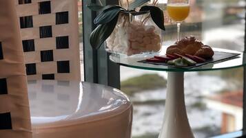 Una habitación moderna con una silla blanca, una mesa de vidrio con un jarrón de flores blancas, un vaso de jugo de naranja y un croissant.