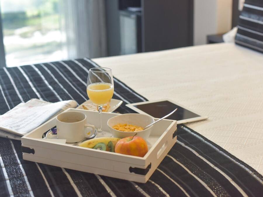 A neatly arranged breakfast tray on a bed with a striped pillow, a smartphone, a glass of orange juice, a bowl of cereal, and a cup of coffee.