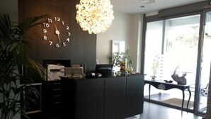 A modern reception desk with a clock, a printer, and a decorative plant. - Hotel Noia (Noia)
