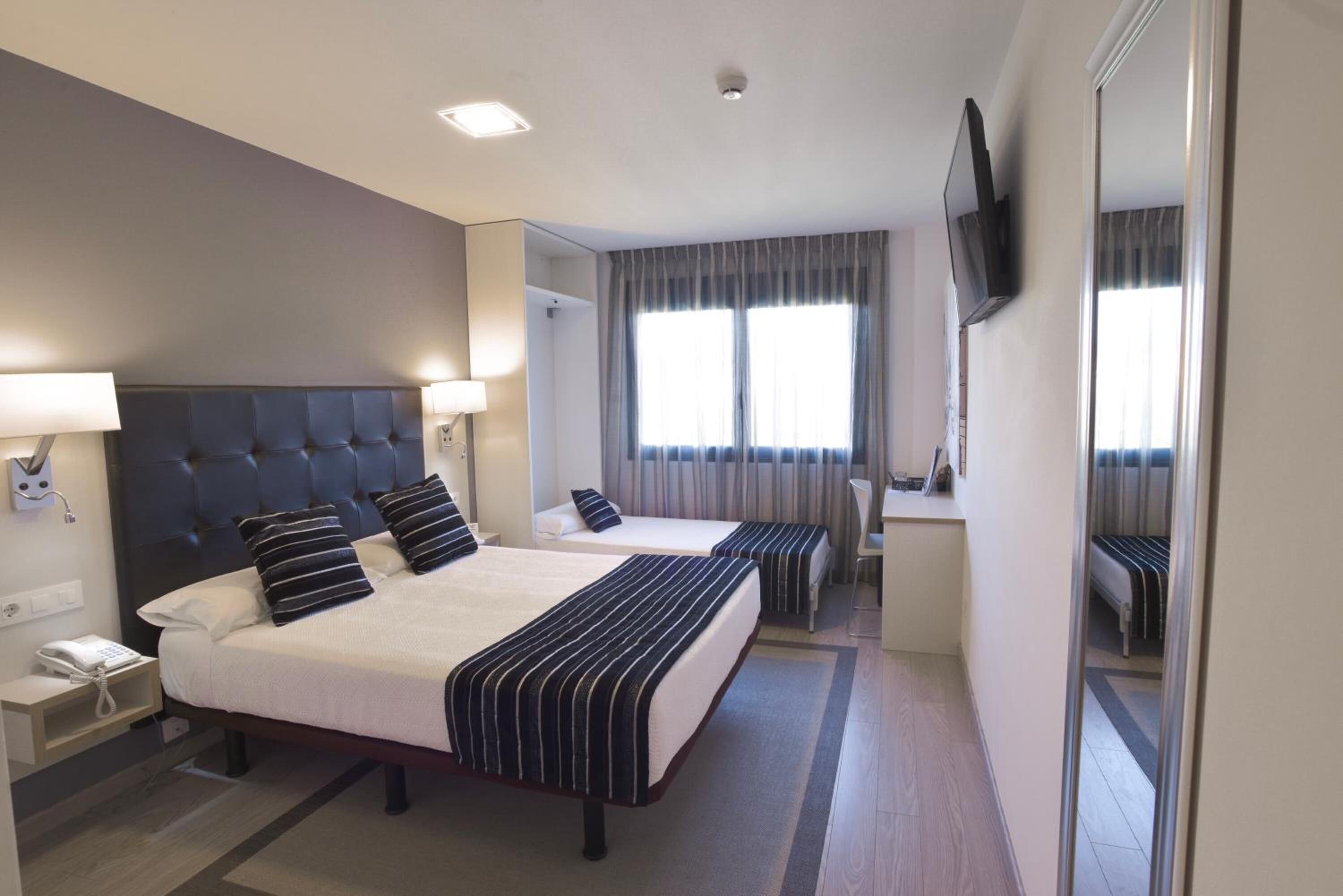 A hotel room with two beds, a desk, a chair, a TV mounted on the wall, and a telephone on the nightstand.