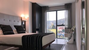 A modern hotel room with a large bed, a bedside table, a lamp, a view of the city through a large window, and a balcony with a chair. - Hotel Noia (Noia)