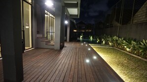 A wooden deck with steps leading to a building, surrounded by a well-lit garden area. - M Boutique Hostel (Keroboken Kelod)