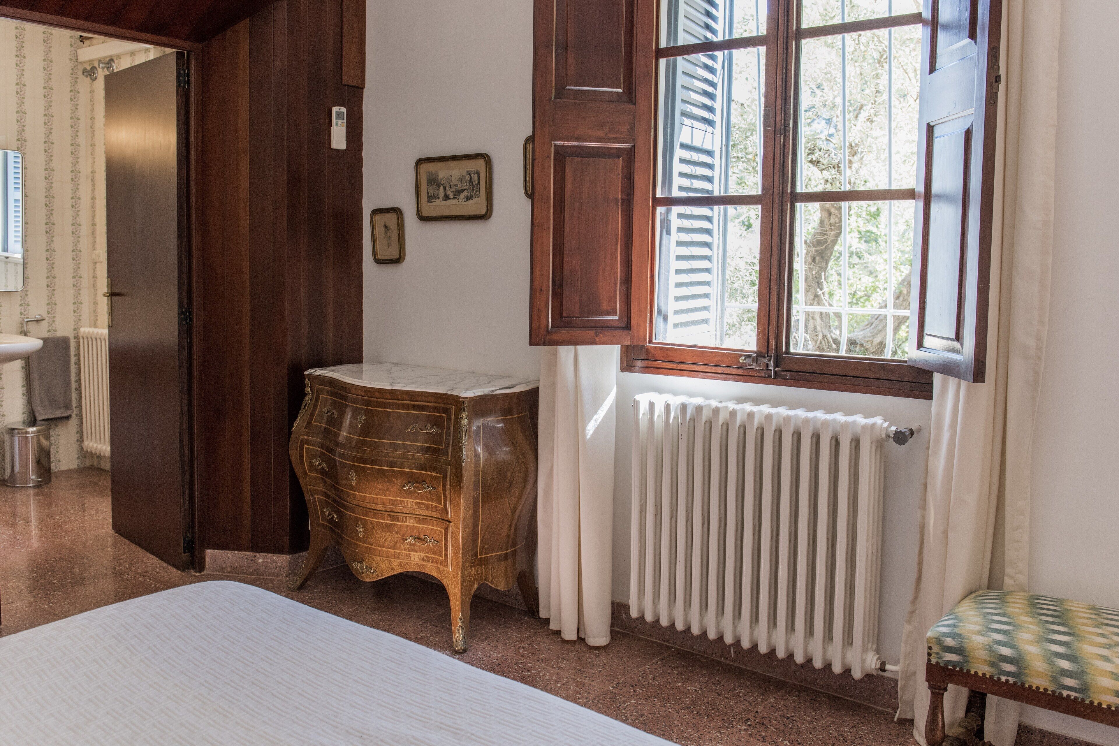 A bedroom with a wooden dresser, a radiator, a window with shutters, and a bench.